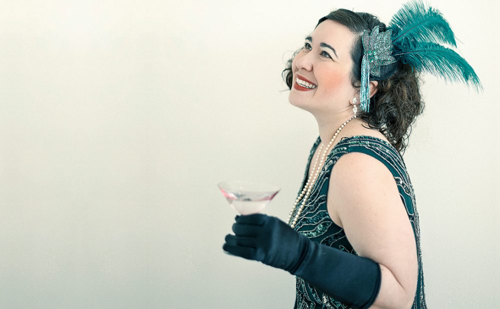 A woman in a green sleeveless dress, with elbow-length gloves and a feathered hair piece, drinking from a coup.