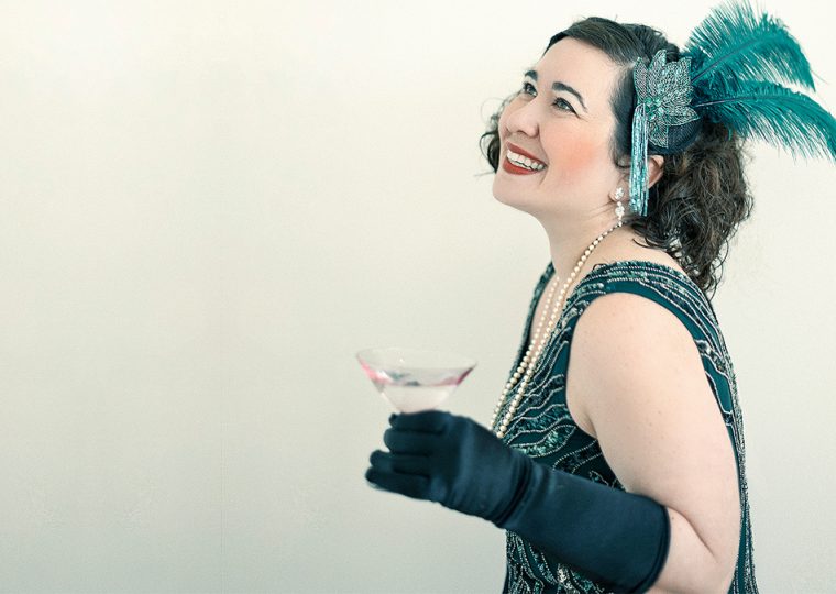 A woman in a green sleeveless dress, with elbow-length gloves and a feathered hair piece, drinking from a coup.