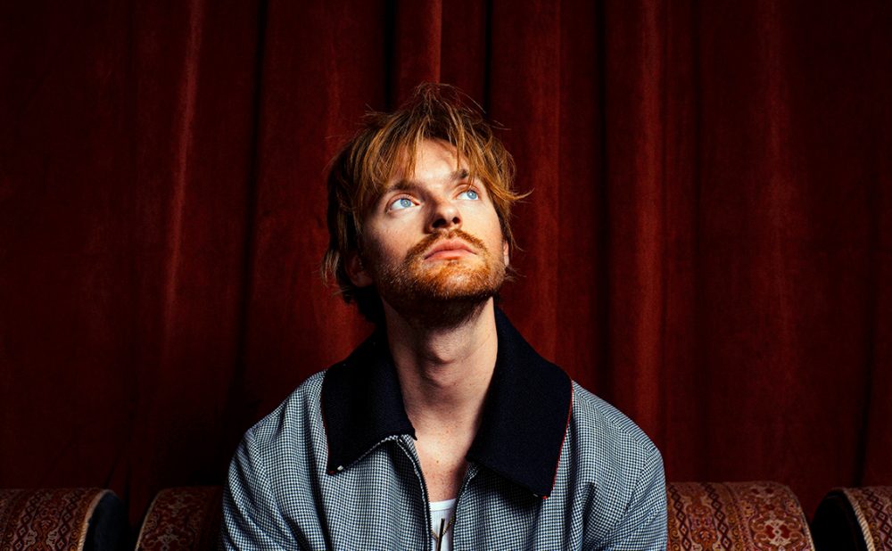 Finneas sitting in front of red velvet drapes, looking up.