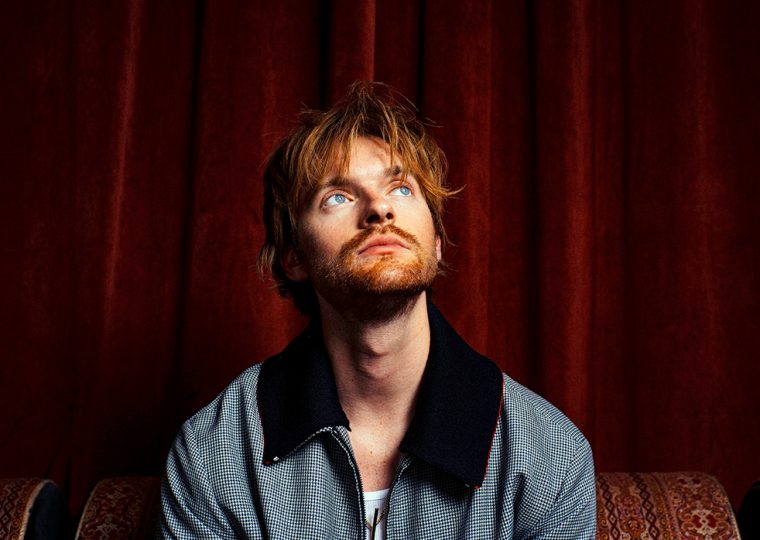 Finneas sitting in front of red velvet drapes, looking up.