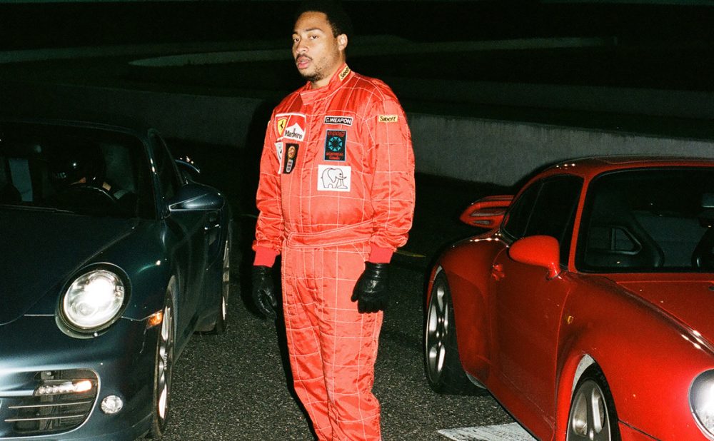 Cadence Weapon standing between two sports cars.