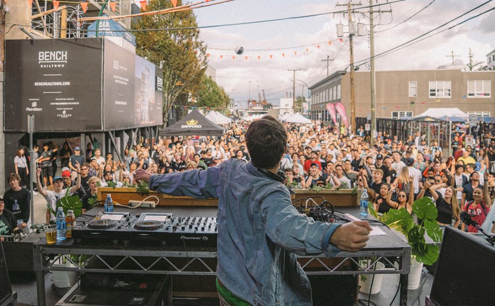 View standing behind a DJ with a crowd of people in front of the DJ booth.
