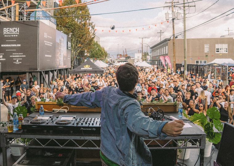 View standing behind a DJ with a crowd of people in front of the DJ booth.