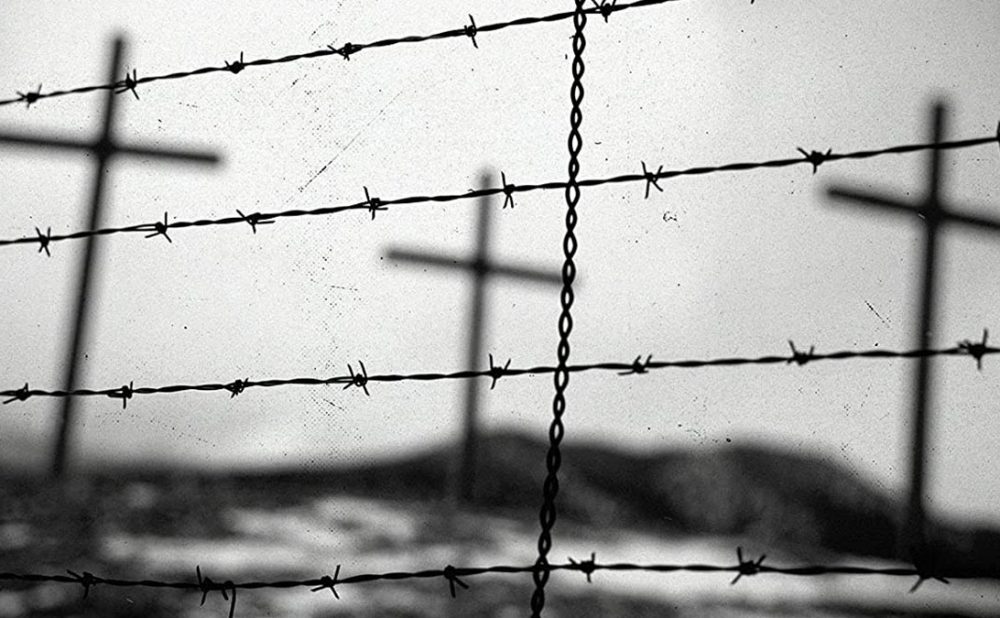 Black and white photo of three crosses in a field, seen behind a barbed wire fence.