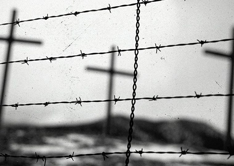 Black and white photo of three crosses in a field, seen behind a barbed wire fence.