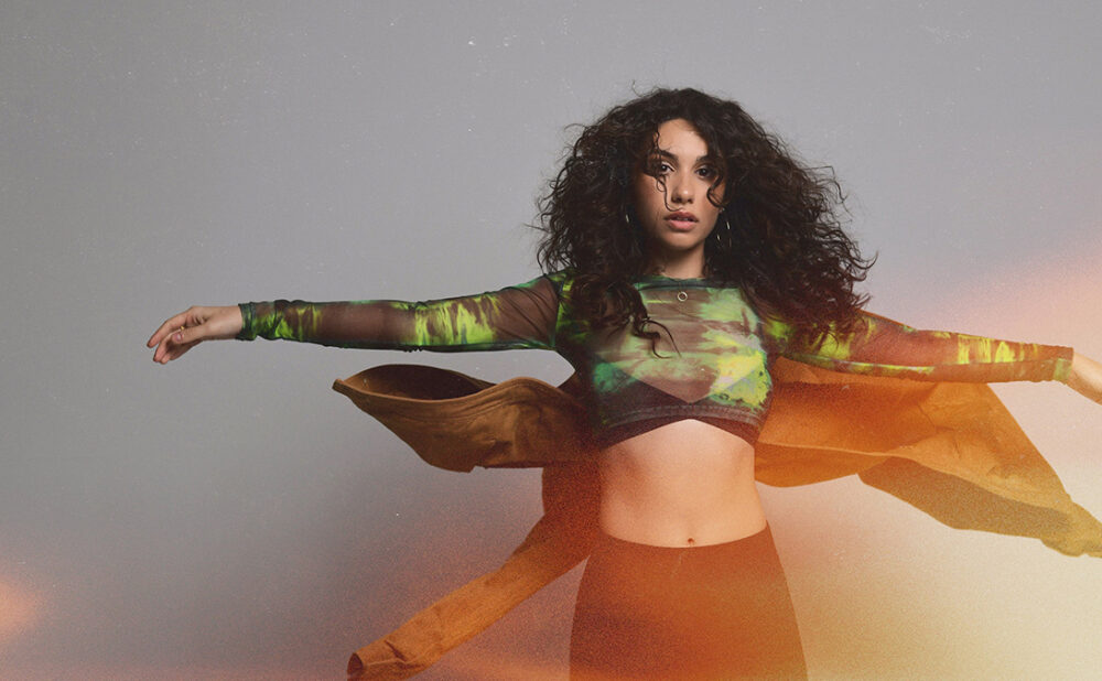 Alessia Cara with her arms held out to each side, as if she's mid-spin, against a grey backdrop