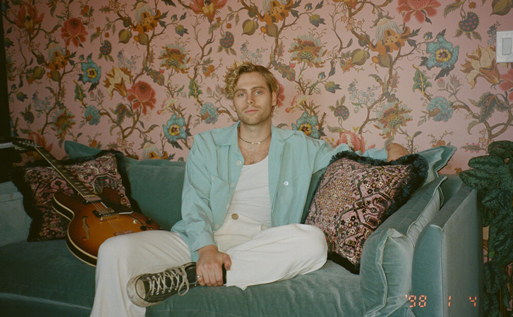 Luke Hemmings casually sitting on a soft teal velvet sofa against a floral wallpapered wall.