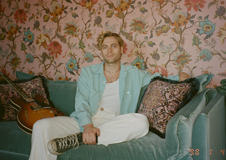 Luke Hemmings casually sitting on a soft teal velvet sofa against a floral wallpapered wall.