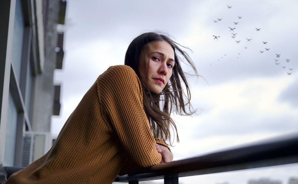 A still from the movie Night Raiders of a women in a camel coloured long sleeve shirt, leaning on the railing of a balcony with a flock of birds flying in the sky