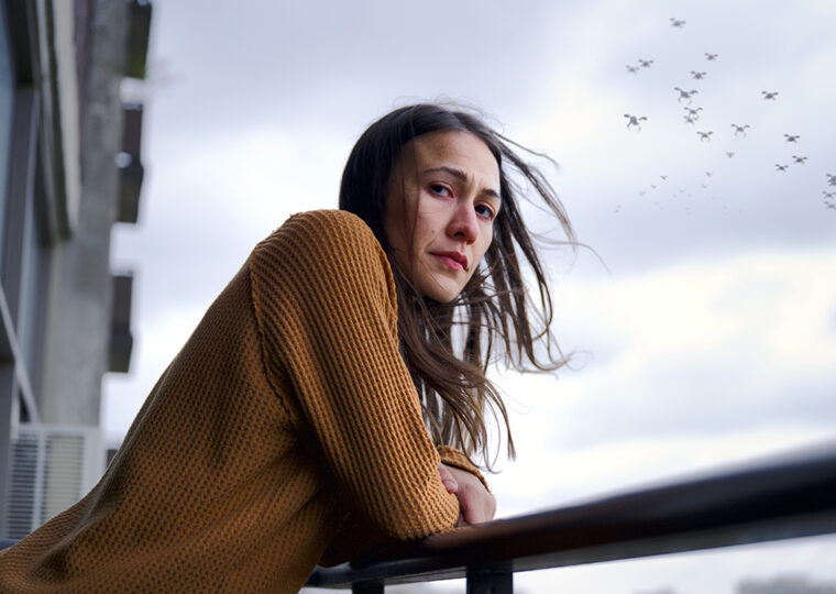 A still from the movie Night Raiders of a women in a camel coloured long sleeve shirt, leaning on the railing of a balcony with a flock of birds flying in the sky