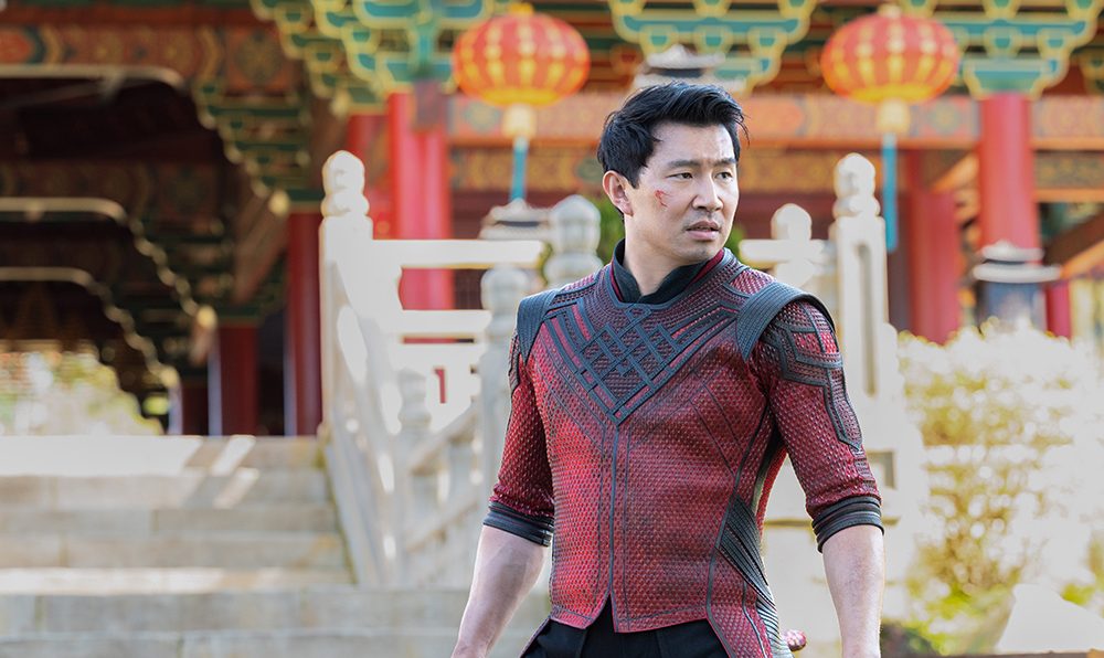 Simu Liu in a black and red suit holding a stave by his side.