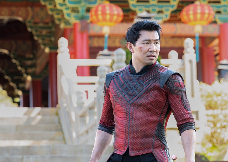 Simu Liu in a black and red suit holding a stave by his side.