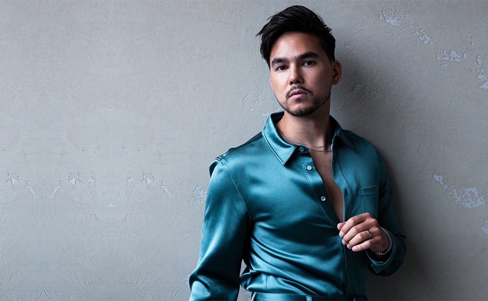 Tyler Shaw standing against a grey wall wearing a teal dress shirt.