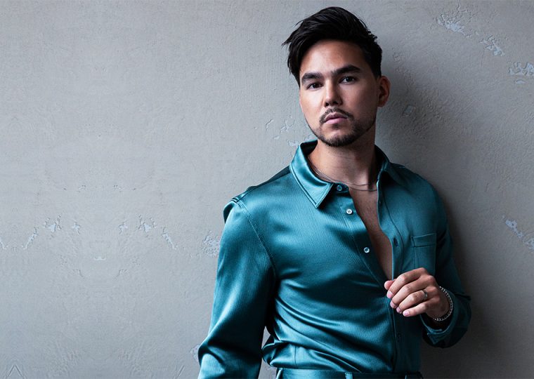 Tyler Shaw standing against a grey wall wearing a teal dress shirt.