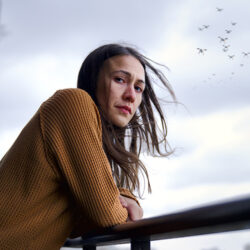 A still from the movie Night Raiders of a women in a camel coloured long sleeve shirt, leaning on the railing of a balcony with a flock of birds flying in the sky