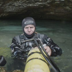 A man in a diver wetsuit, standing in waist-high water, holding a yellow oxygen tank.