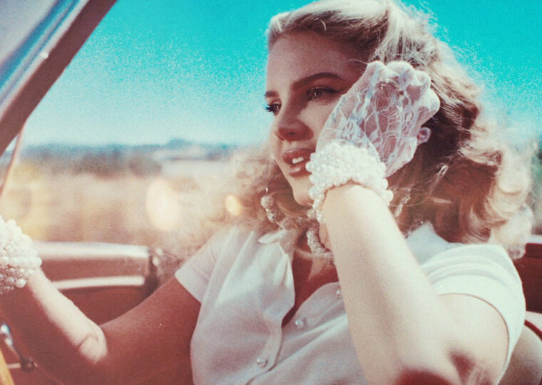 Lana Del Ray driving a convertible, dressed in white wearing white lace gloves.