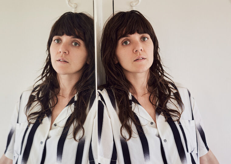 Courtney Barnett leaning against a full length mirror in a black and white top.