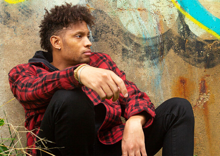 Jhyve sitting against a graffiti'd wall in a red plaid jacket and black pants.