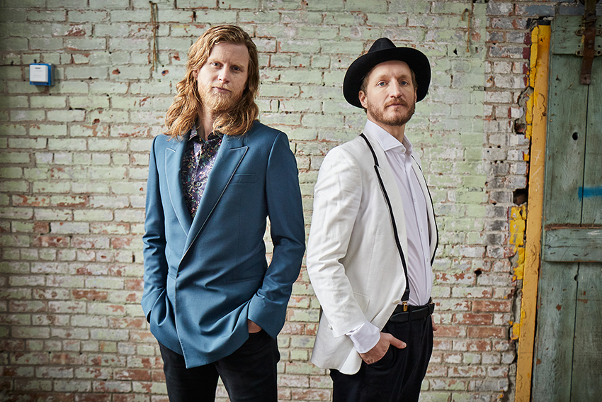 Wesley Schultz (left) and Jeremiah Fraites (right) standing facing slightly away from each other with a white washed brick wall behind them.