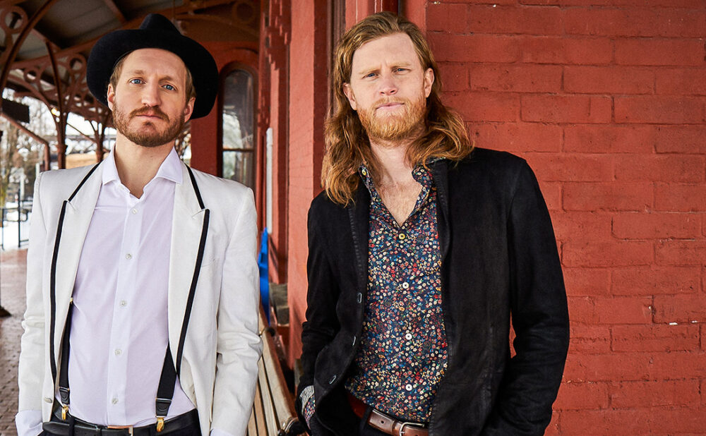 Jeremiah Fraites (left) and Wesley Schultz from The Lumineers standing side by side in front of a painted red brick wall.
