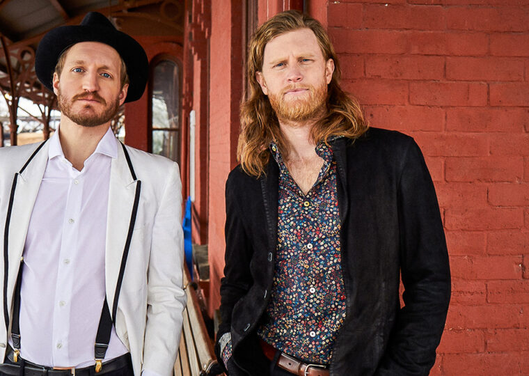 Jeremiah Fraites (left) and Wesley Schultz from The Lumineers standing side by side in front of a painted red brick wall.