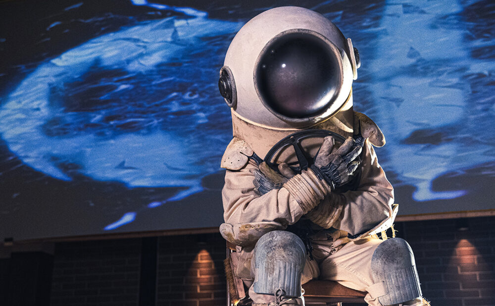 Scene from the film Do You Mind If I Sit Here? of someone wearing an old diver suit clutching a film reel, sitting on a chair.