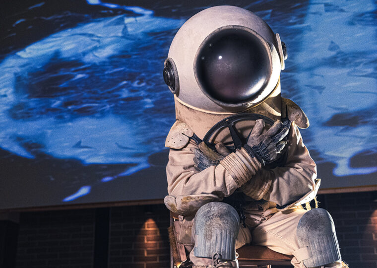 Scene from the film Do You Mind If I Sit Here? of someone wearing an old diver suit clutching a film reel, sitting on a chair.