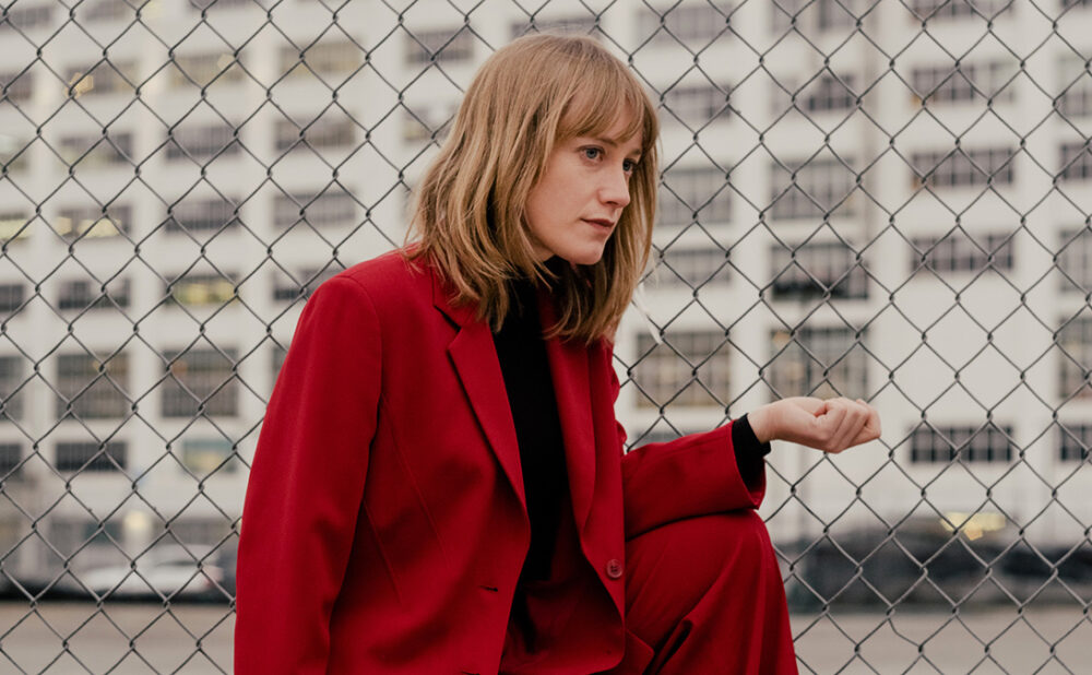 Photo of Tamara Lindeman dressed in a red pant suit, crouching outside next to a wire fence.