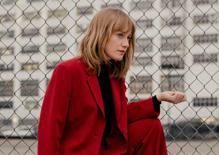 Photo of Tamara Lindeman dressed in a red pant suit, crouching outside next to a wire fence.