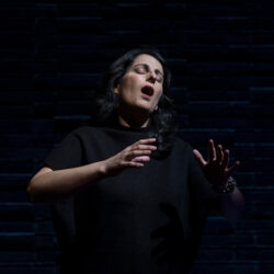 Zorana Sadiq dressed in all black against a black backdrop singing on stage.