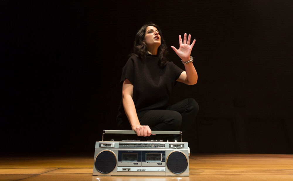 Zorana Sadiq on stage dressed in all black against a black backdrop, her hand holding a silver boombox.