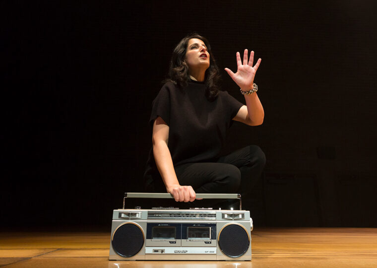 Zorana Sadiq on stage dressed in all black against a black backdrop, her hand holding a silver boombox.
