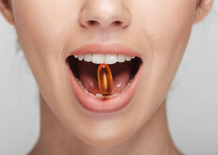 A stock photo of a mouth holding a orange capsule between their teeth.