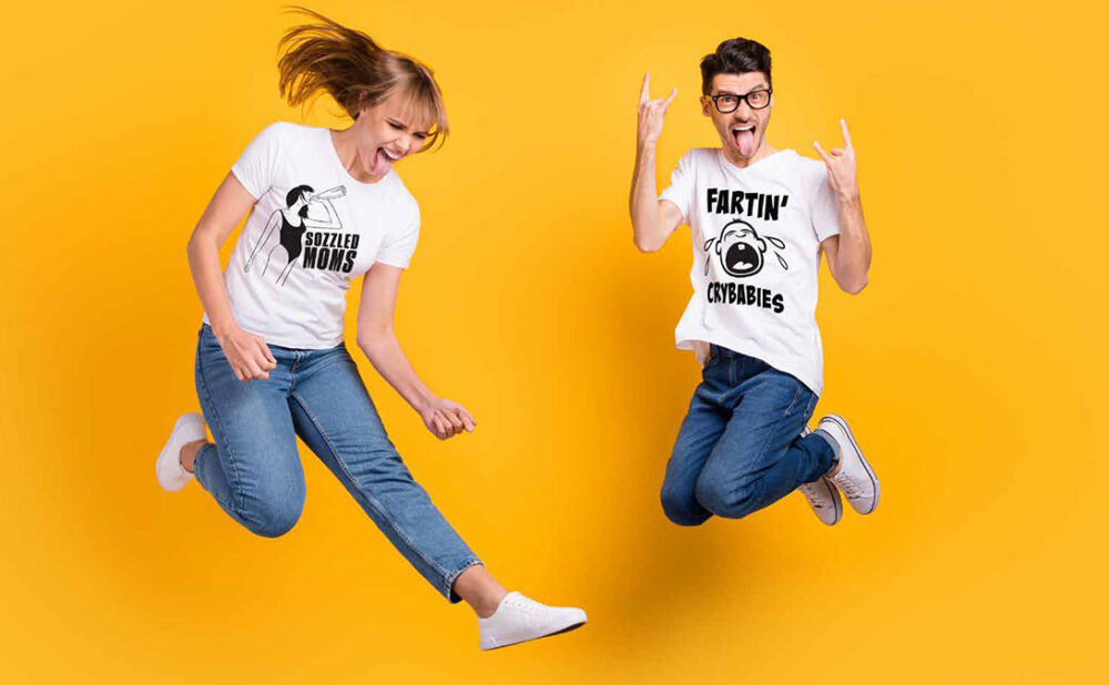 A woman and a man wearing band t-shirts and blue jeans jumping in the air against a yellow backdrop.