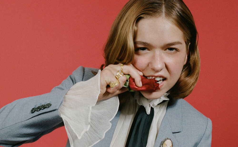 Lindsay Jordan (Snail Mail) biting a rose, dressed in grey suit and white blouse against a red-orange backdrop.