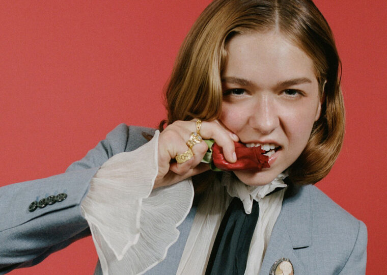 Lindsay Jordan (Snail Mail) biting a rose, dressed in grey suit and white blouse against a red-orange backdrop.