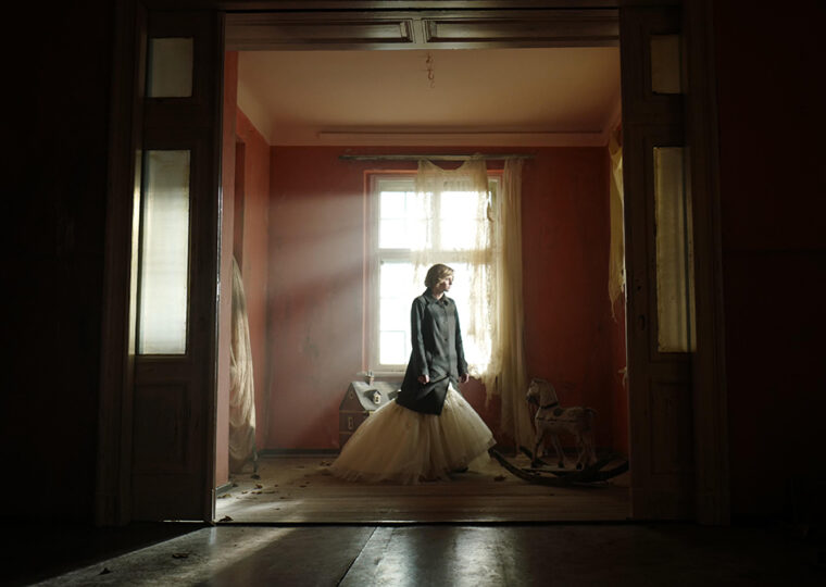 Scene from the film Spencer of Princess Di, played by Kristen Stewart, standing in a nearly bare room in front of a window with the sun streaming in.
