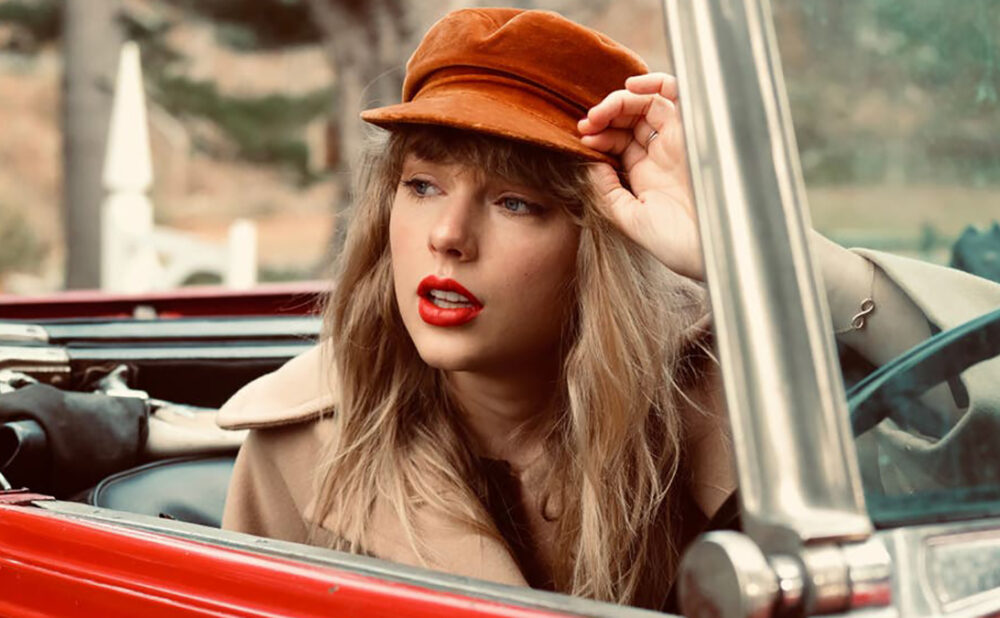 Taylor Swift sitting in a red convertible, top down, wearing a orange-brown hat and camel coloured coat.