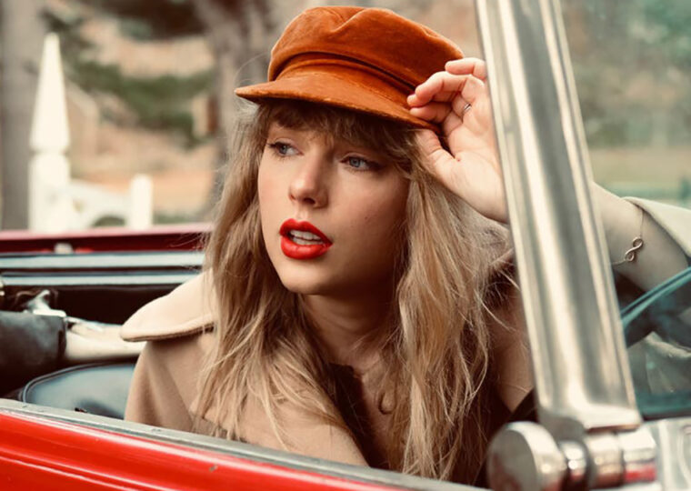 Taylor Swift sitting in a red convertible, top down, wearing a orange-brown hat and camel coloured coat.
