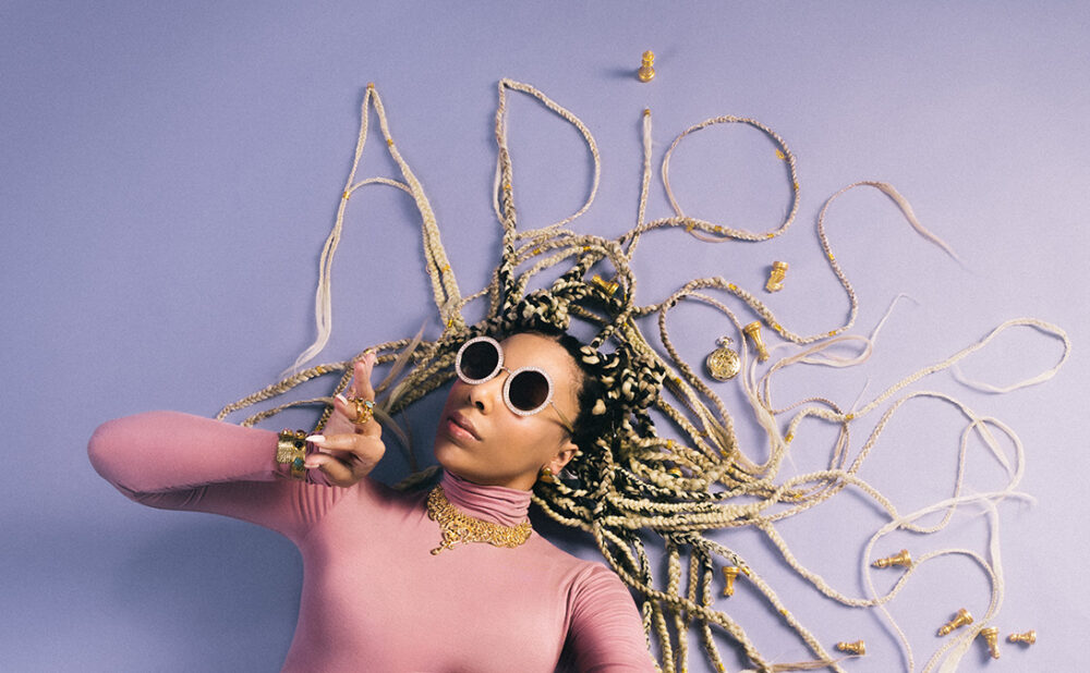 Odreii lying on a lavender coloured floor wearing sunglasses, with her hair shaped to spell out