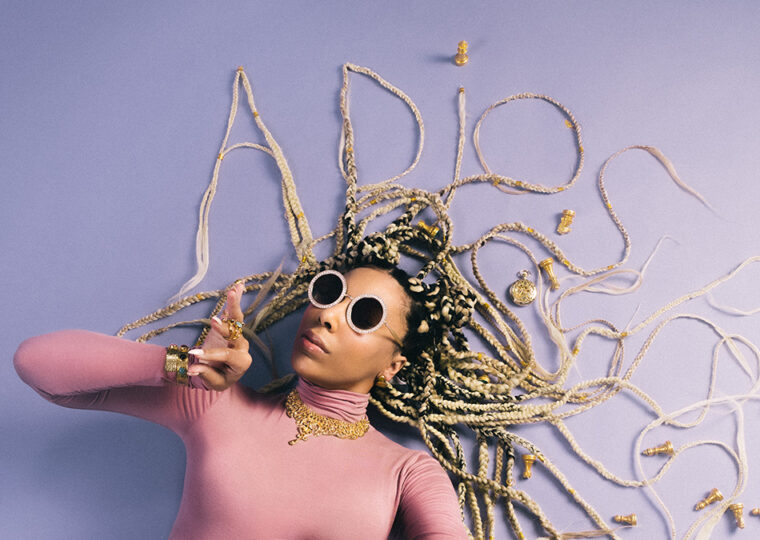 Odreii lying on a lavender coloured floor wearing sunglasses, with her hair shaped to spell out 