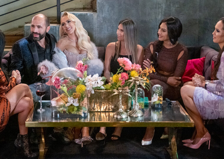 Cast of Selling Sunset Season 4 talking with each other, sitting around a coffee table topped with flowers and drinks.